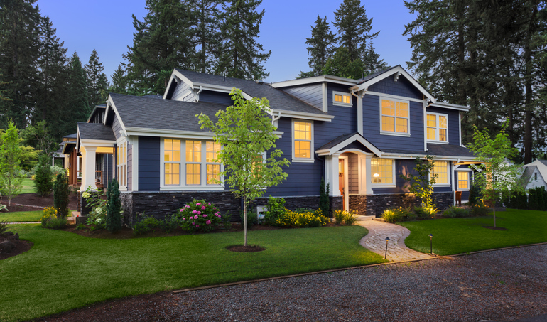 facade of home with manicured lawn, landscaping, and backdrop of trees and dark blue sky. Glowing interior lights create a welcoming mood.