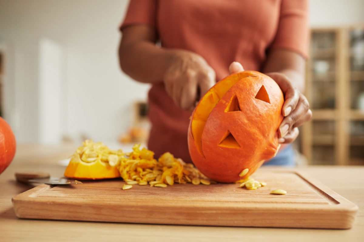 Unrecognizable African American woman standing at table carving Jack-O'-Lantern out of ripe orange pumpkin