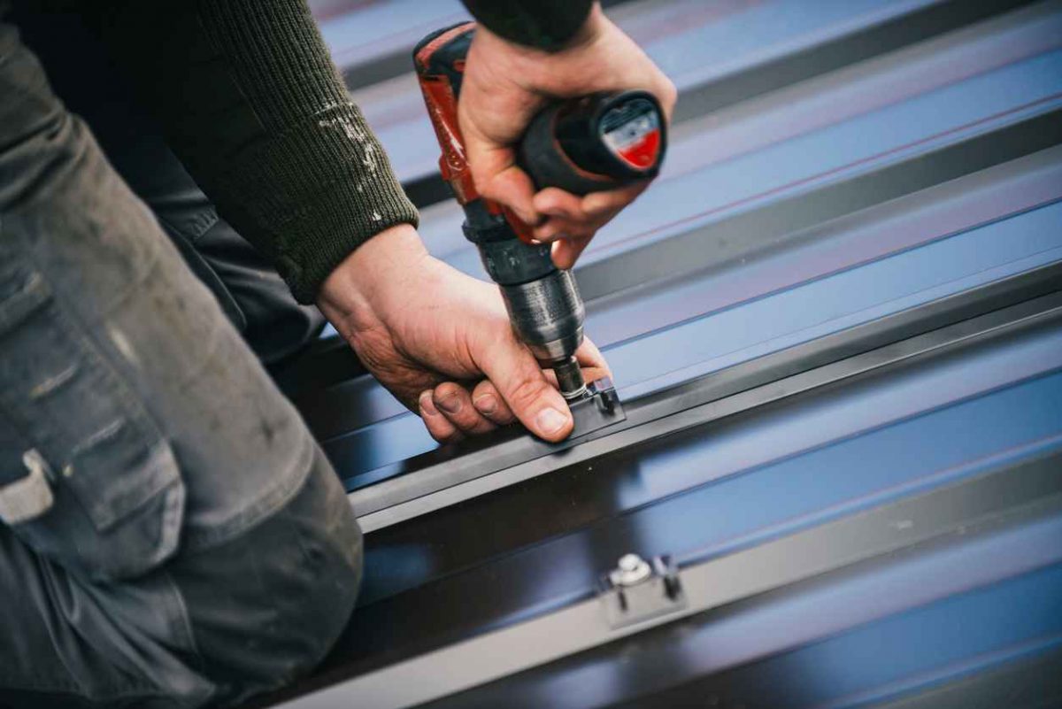 Building contractor is installing metal roofing sheets on the rooftop of the house using electric screwdriver.
